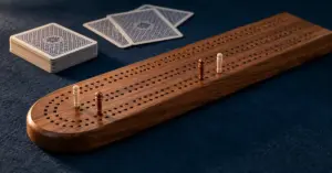 Puinen cribbage-lauta, vaaleat ja kuparinväriset pistetikut sekä pelikortit tummansinisellä pelipöydällä.